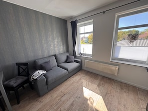 Un salon moderne avec un canapé gris, un fauteuil noir et une fenêtre donnant sur l’extérieur.