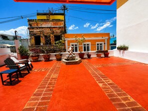 Eine Dachterrasse mit rotem Boden, Kübelpflanzen, einer Statue und einem Gebäude mit Balkonen im Hintergrund.