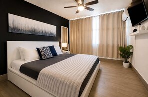 Ein Schlafzimmer mit einem Bett, einem Deckenventilator, einem Fernseher und einem Fenster mit Vorhängen.