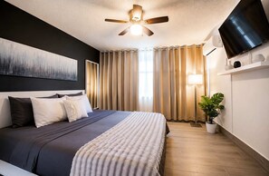 Quarto de hotel moderno com cama, ventilador de teto, TV de tela plana, planta em vaso e uma grande janela com cortinas.