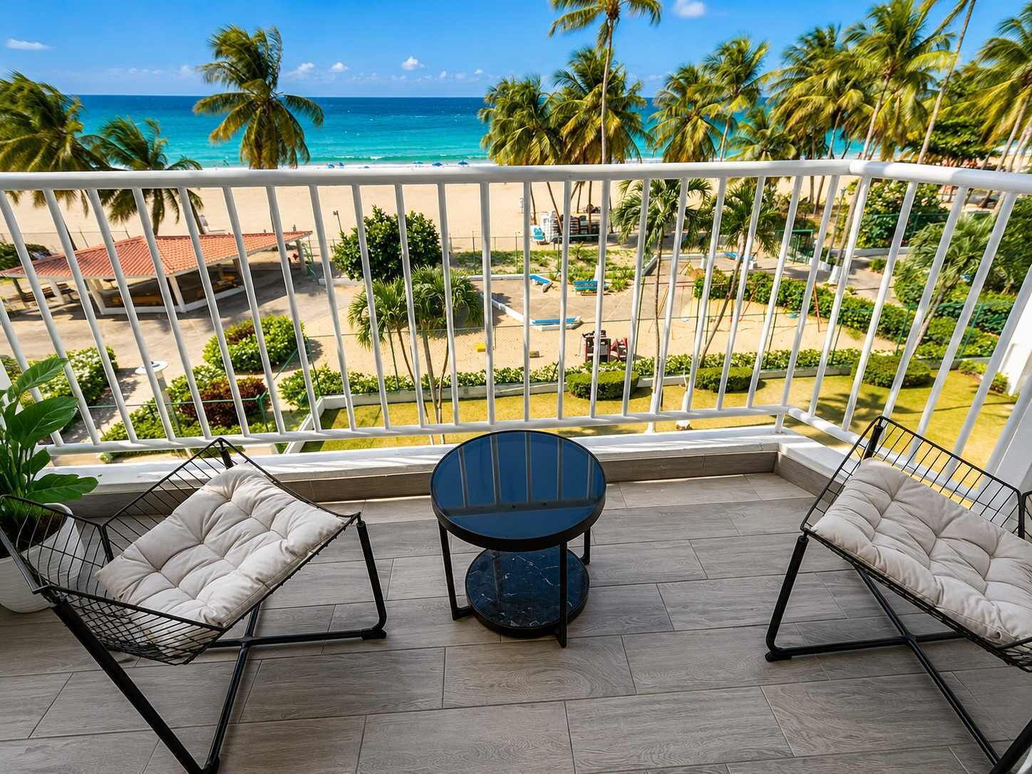 A balcony with two cushioned chairs, a small round table, and a view of a beach with palm trees and a pool.