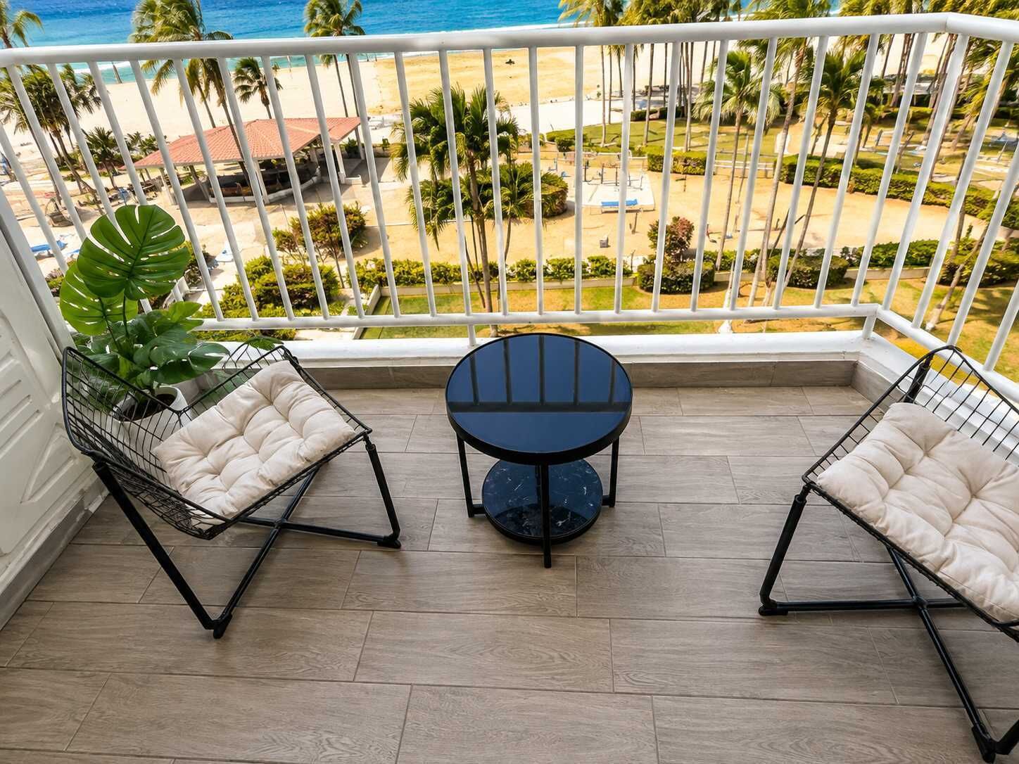 A balcony with two wire-framed chairs and a small round table, overlooking a beach and palm trees.
