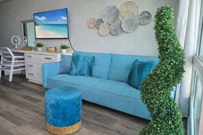 A living room with a blue sofa, a round ottoman, a desk with a chair, and a wall with decorative plates. - ESJ Towers Studio - Isla Verde Beach Access (Avenida Isla)