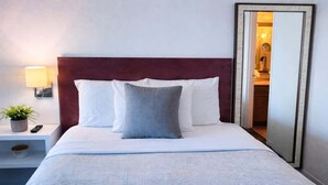 Ein ordentlich bezogenes Bett mit weißen und grauen Kissen, einem Holzkopfende, einer Nachttischlampe, einer kleinen Pflanze und einem Spiegel an der Wand.