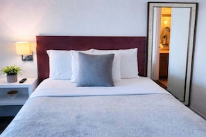 A neatly made bed with white and gray pillows, a wooden headboard, a bedside lamp, a small plant, and a mirror on the wall. - ESJ Towers Studio - Isla Verde Beach Access (Avenida Isla)