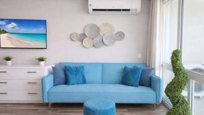 Ein Wohnzimmer mit einem blauen Sofa, einem runden Hocker, einer Wand-Klimaanlage, einem Strand-Gemälde, dekorativen Tellern und einer Topfpflanze.