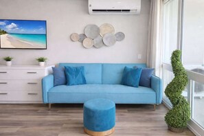 A living room with a blue sofa, a round ottoman, a wall-mounted air conditioner, a beach-themed painting, decorative plates, and a potted plant. - ESJ Towers Studio - Isla Verde Beach Access (Avenida Isla)