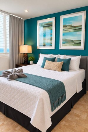 A hotel room with a bed, a nightstand, a lamp, a window with curtains, and two framed pictures on the wall. - Queen Bed in 1BR Beachfront Apt Condado San Juan (SanJuan)