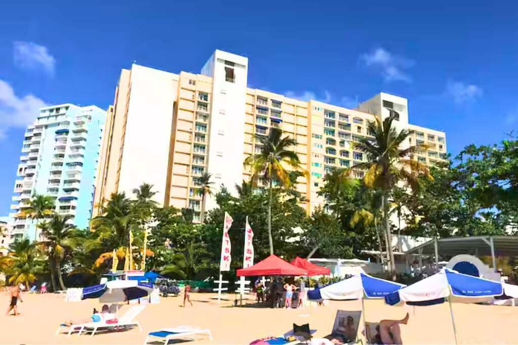 Um hotel à beira-mar com vários andares, palmeiras e guarda-sóis de praia.