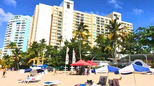 Un hotel frente a la playa con varios pisos, palmeras y sombrillas.