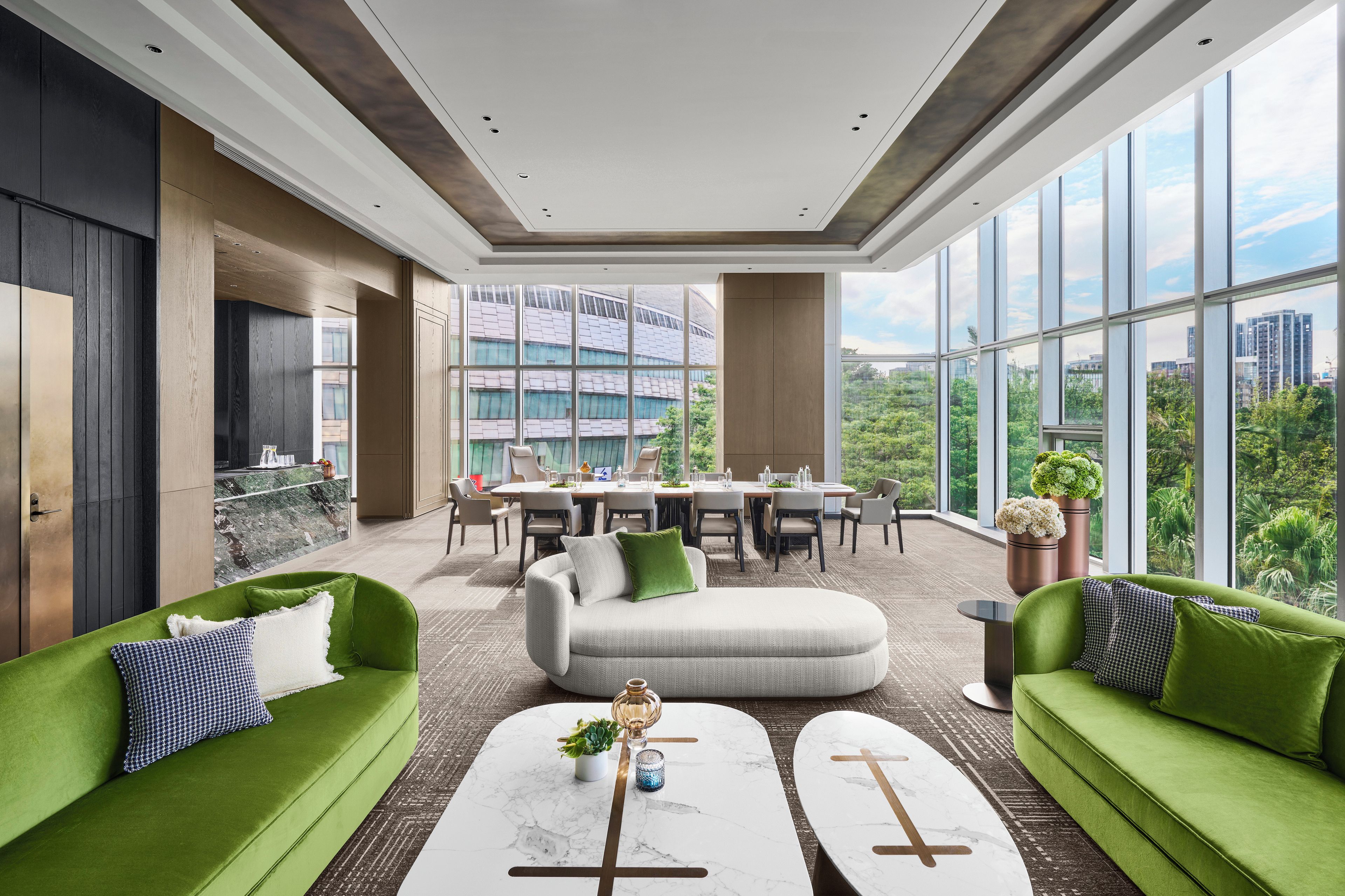 A modern living room with green sofas, a marble coffee table, and a dining area with a view of a cityscape through large windows.