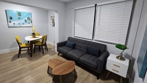 A modern living room with a dark grey sofa, a round wooden coffee table, a white side table with a green lamp, and a painting of palm trees on the wall. - The Sunny 2-Bedroom Retreat | POOL (Miami)