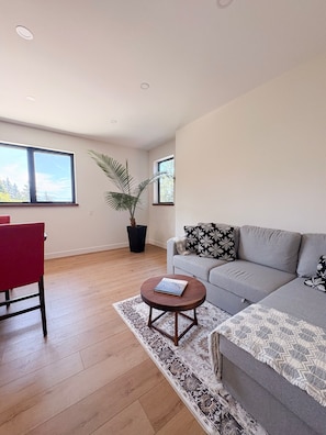 A modern living room with a grey sofa, a red chair, a round wooden coffee table, a patterned rug, and a large window with a view of trees.