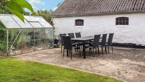 Een kas met glazen constructie, een wit gebouw met pannendak en een tafel met stoelen op de voorgrond.