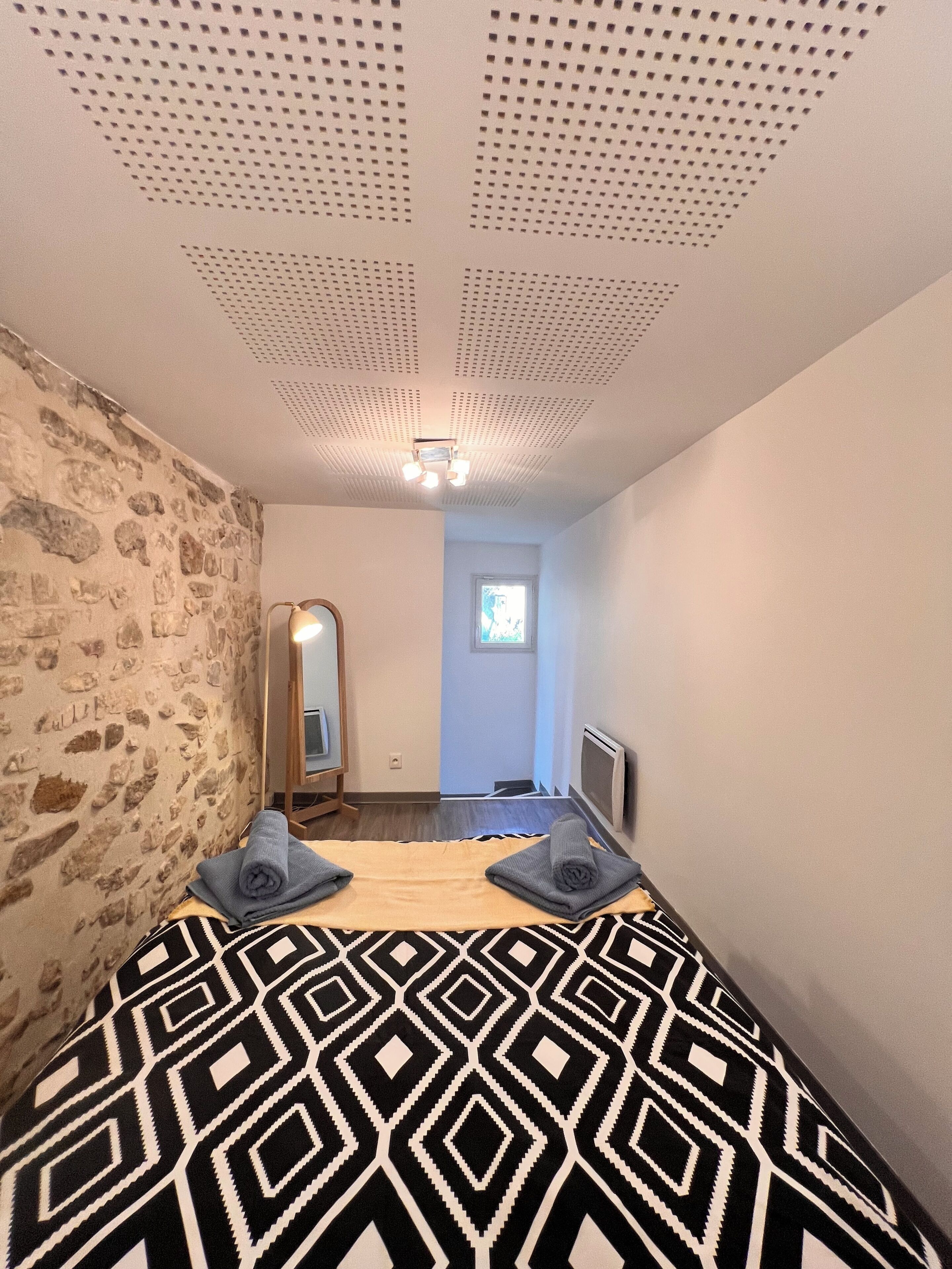A bedroom with a patterned bedspread, a stone wall, a mirror, and a ceiling with a light fixture.