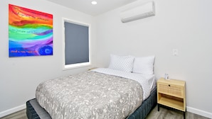 Een slaapkamer met een bed, een nachtkastje, een wandairconditioner, een raam en een kleurrijk poster.