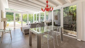 Een modern interieur met een glazen eettafel, transparante stoelen, een rode kroonluchter en grote ramen met uitzicht op de omgeving.