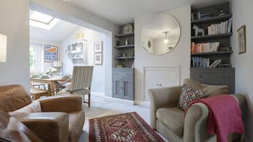 A living room with a sofa, armchair, and a coffee table. There is a bookshelf, a mirror, and a dining area visible in the background.