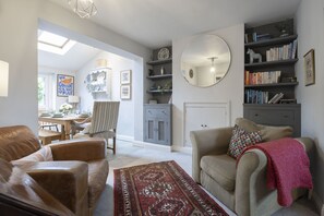 A living room with a sofa, armchair, and a coffee table. There is a bookshelf, a mirror, and a dining area visible in the background. - Charming 3-Bedroom Home with Cosy Log Burner (Cheltenham)