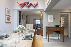 A dining area with a long wooden table set for a meal, a wooden desk with drawers, and a staircase leading to another level. - Charming 3-Bedroom Home with Cosy Log Burner (Cheltenham)