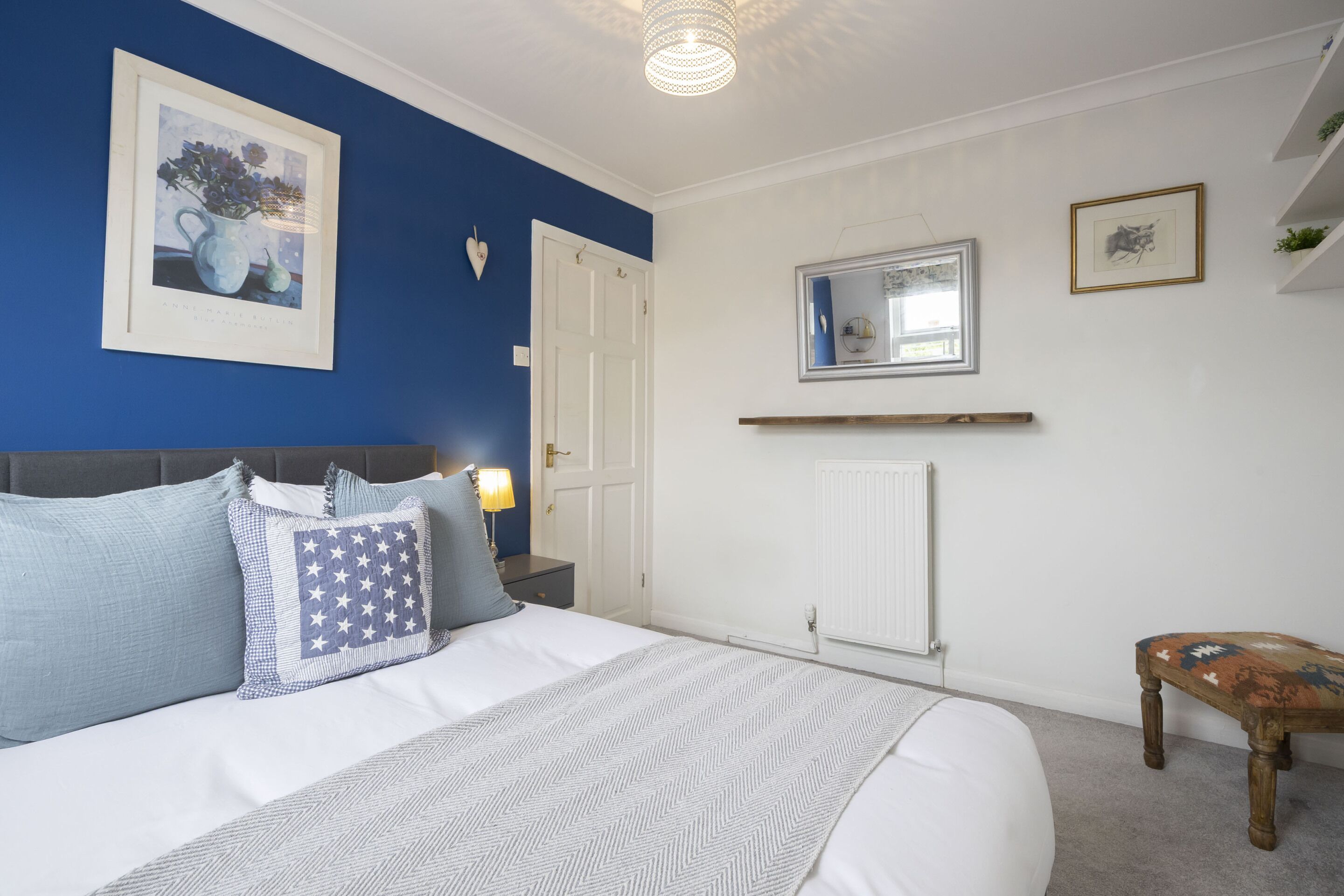 A bedroom with a bed, a nightstand, a lamp, a framed picture, a mirror, and a small wooden table.