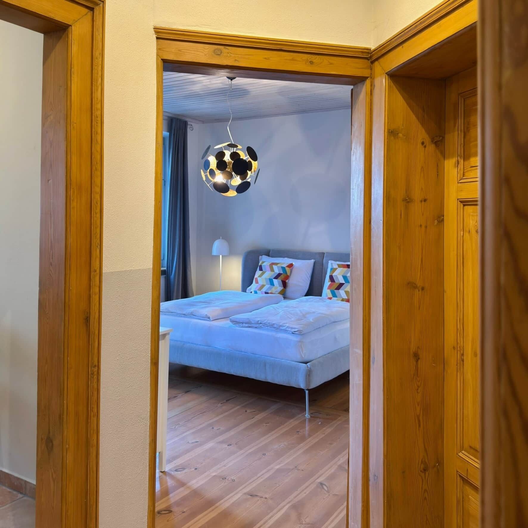 A bedroom with a bed, a hanging light fixture, and wooden flooring.