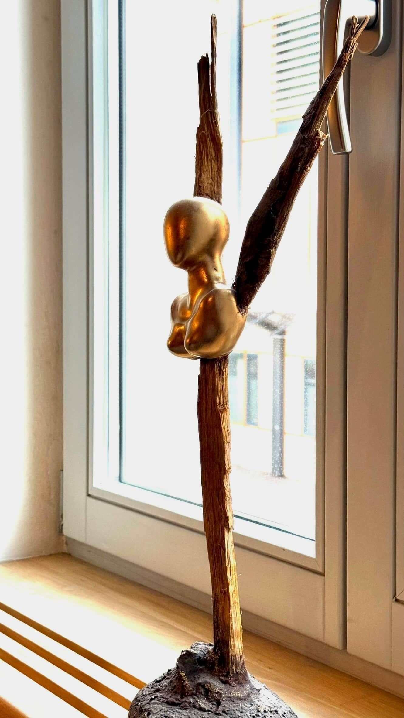 Una escultura de figura humana sobre un pedestal de madera, ubicada cerca de una ventana con persianas.