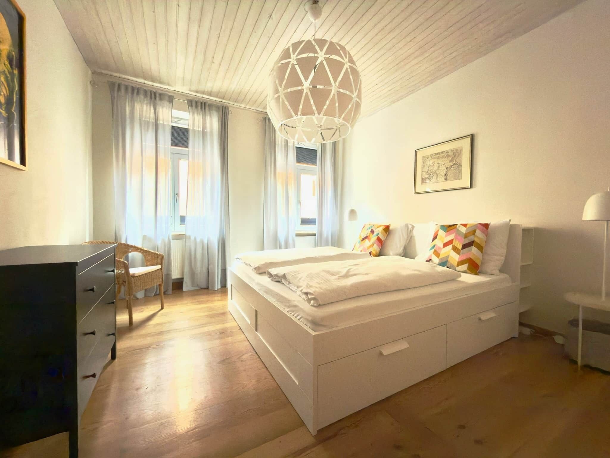 A bedroom with a bed, a desk, a chair, a hanging light fixture, and a window with curtains.