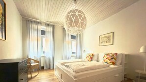 Ein Schlafzimmer mit Bett, Schreibtisch, Stuhl, einer Pendelleuchte und einem Fenster mit Vorhängen.