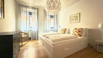 A bedroom with a bed, a desk, a chair, a hanging light fixture, and a window with curtains.