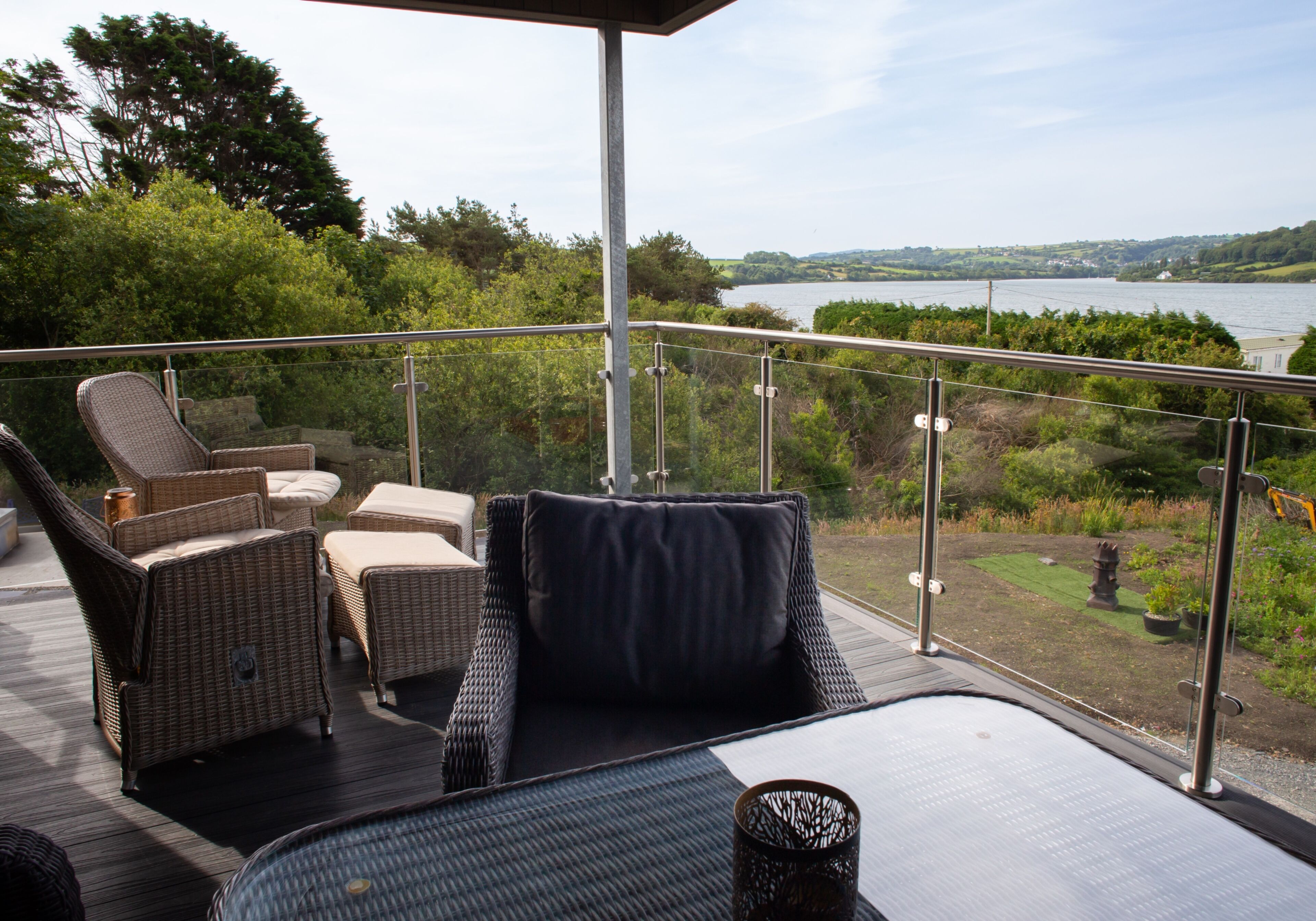 A balcony with wicker furniture, a glass railing, and a view of a lake and greenery.