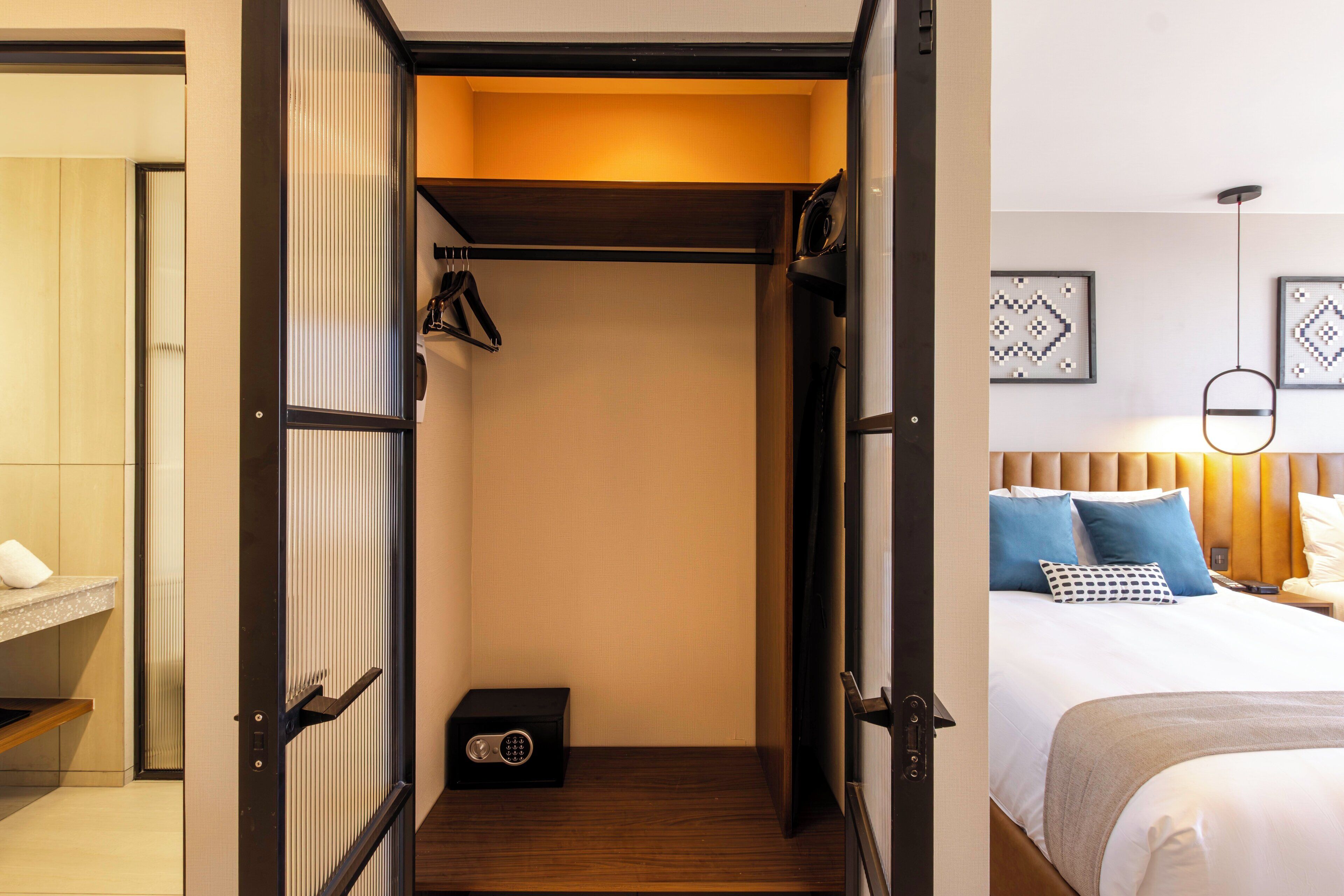 A hotel room with a walk-in closet, a bed with pillows, a bedside table, and a wall-mounted light fixture.