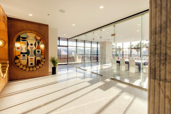 Un hall d’hôtel moderne avec un grand mur de verre, un comptoir d’accueil en bois et une vue sur le paysage urbain.