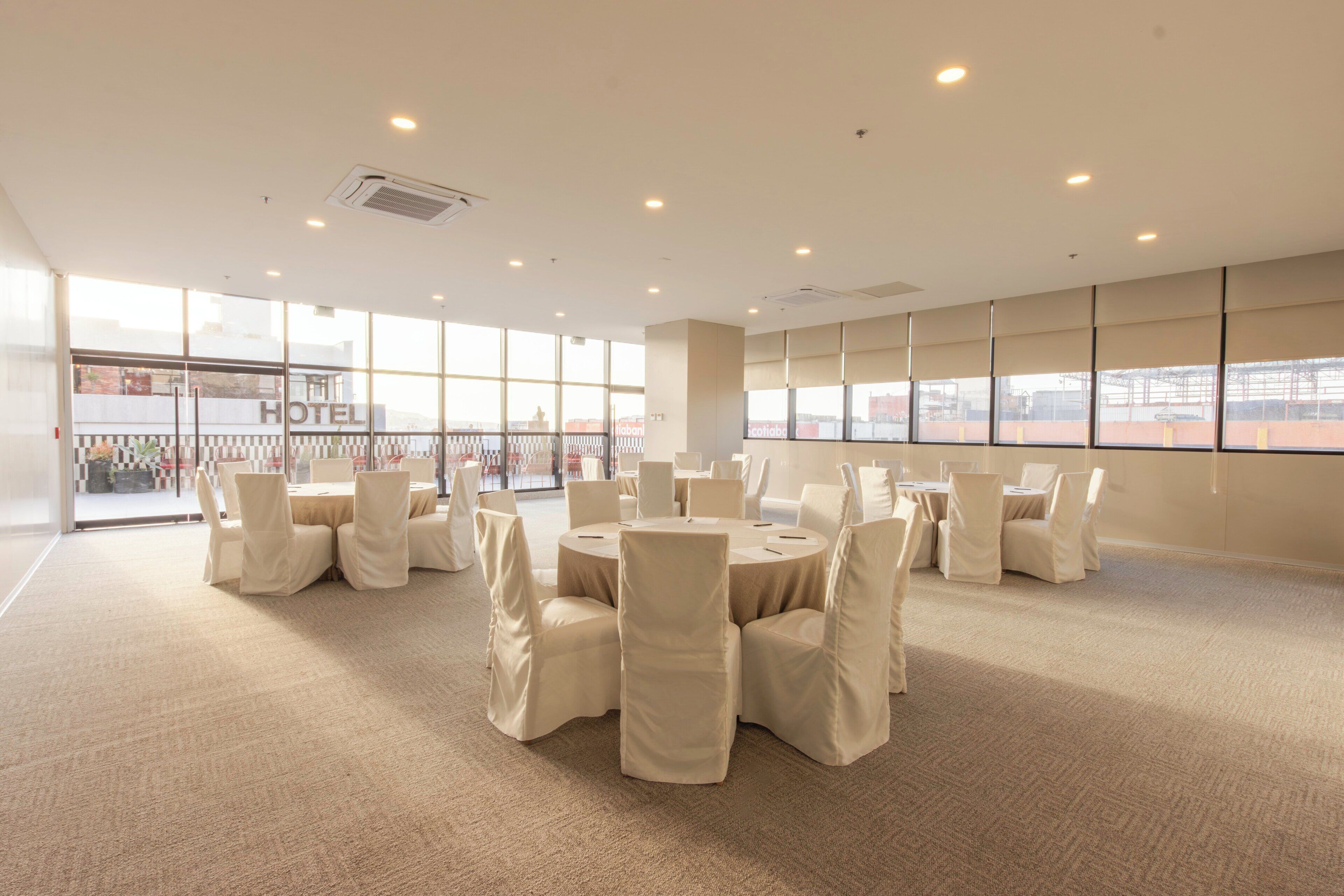 A hotel conference room with round tables, chairs, and large windows.