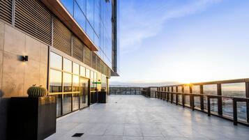 Un edificio moderno con fachada de vidrio, una terraza en la azotea con barandilla y una vista despejada del paisaje urbano a lo lejos.
