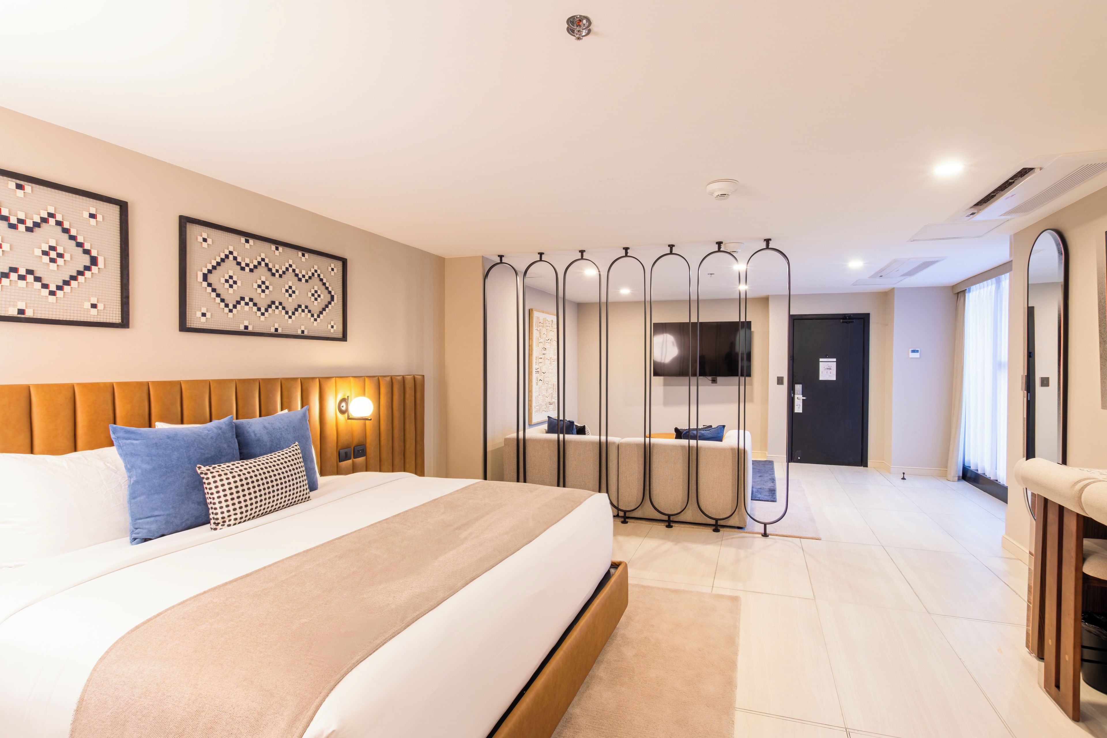 A hotel room with a large bed, a headboard, a bedside table with a lamp, a mirror, and a view of the hallway.