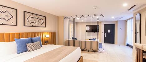 Habitación de hotel con una cama grande, cabecera, mesita de noche con lámpara, espejo y vista al pasillo.