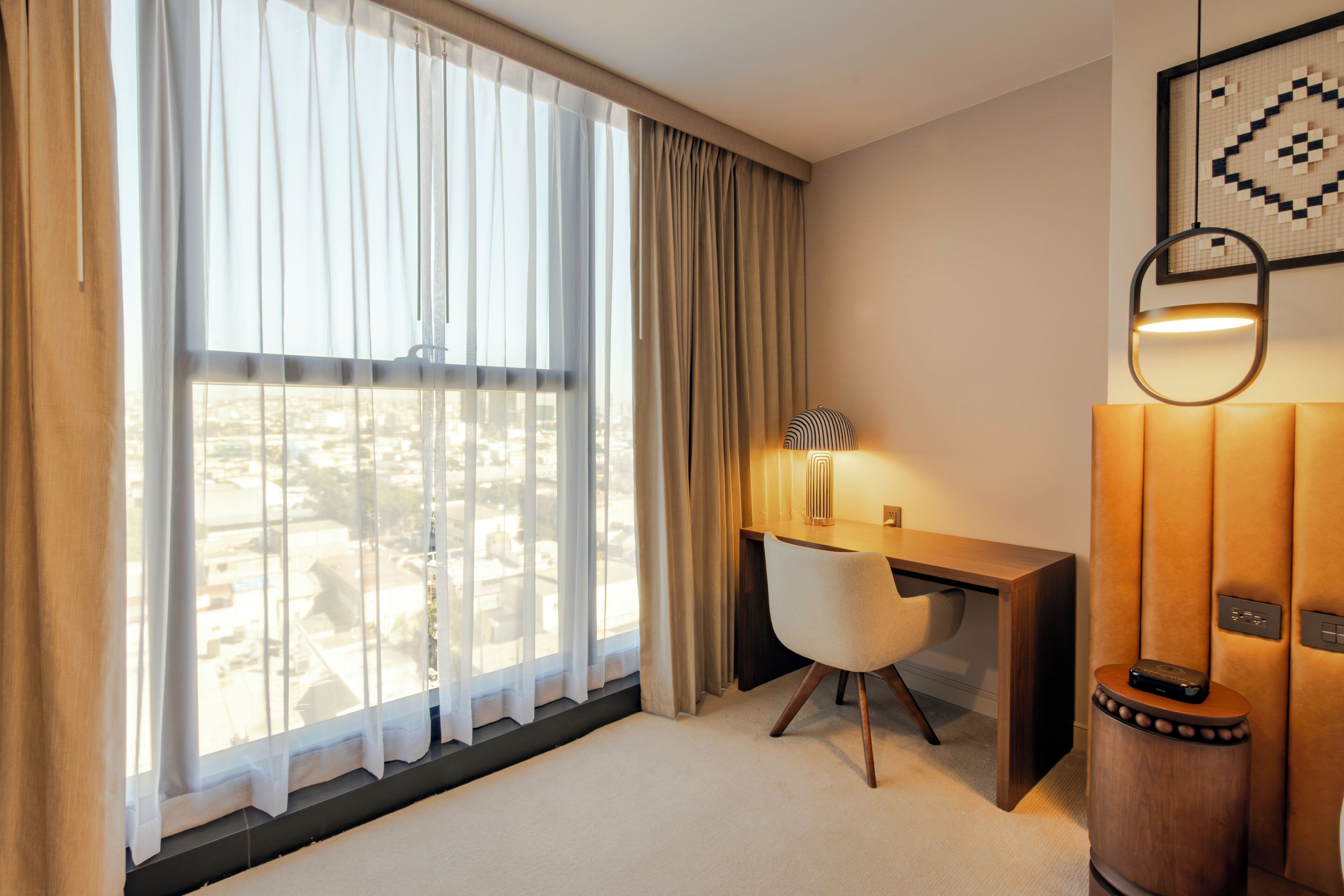 A hotel room with a large window, a desk, a chair, and a wall-mounted lamp.