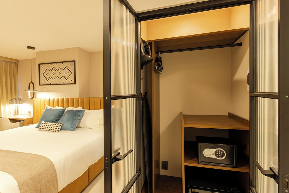 Habitación de hotel con cama, mesita de noche con lámpara, armario con espejo y una caja fuerte.