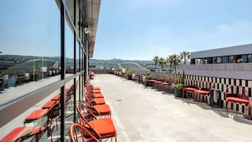 A rooftop terrace with red chairs and tables, overlooking a cityscape with palm trees and buildings.