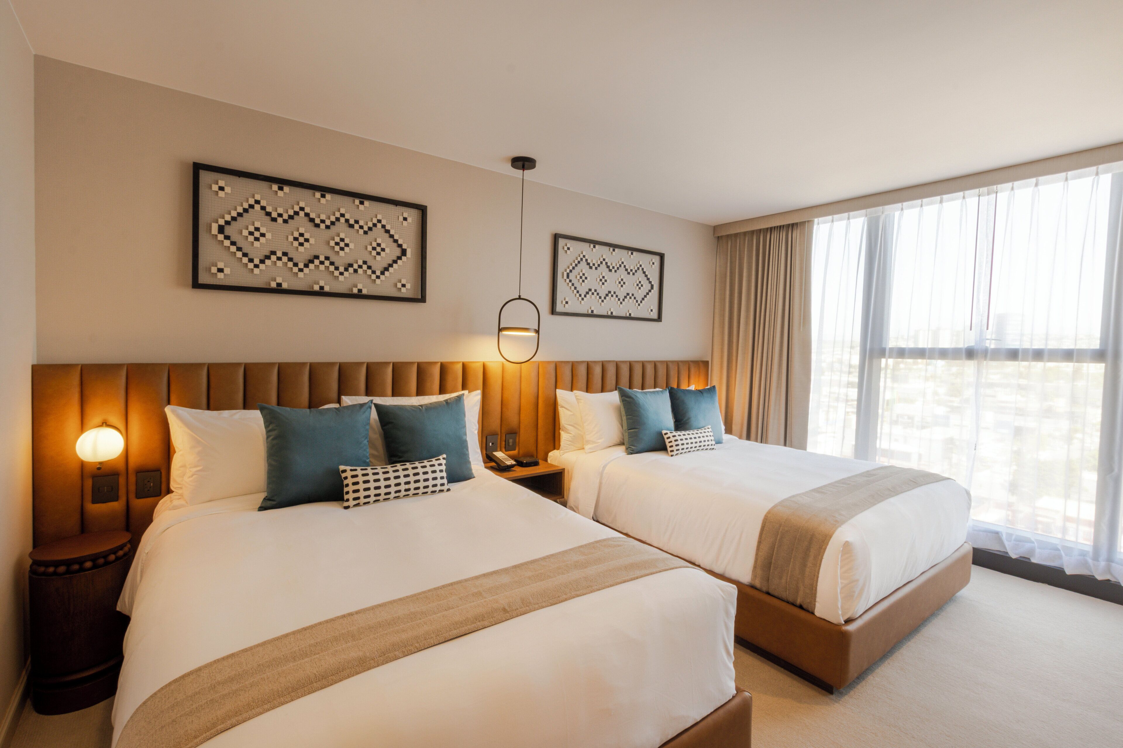 A hotel room with two beds, a headboard, bedside tables, wall art, and a window with curtains.