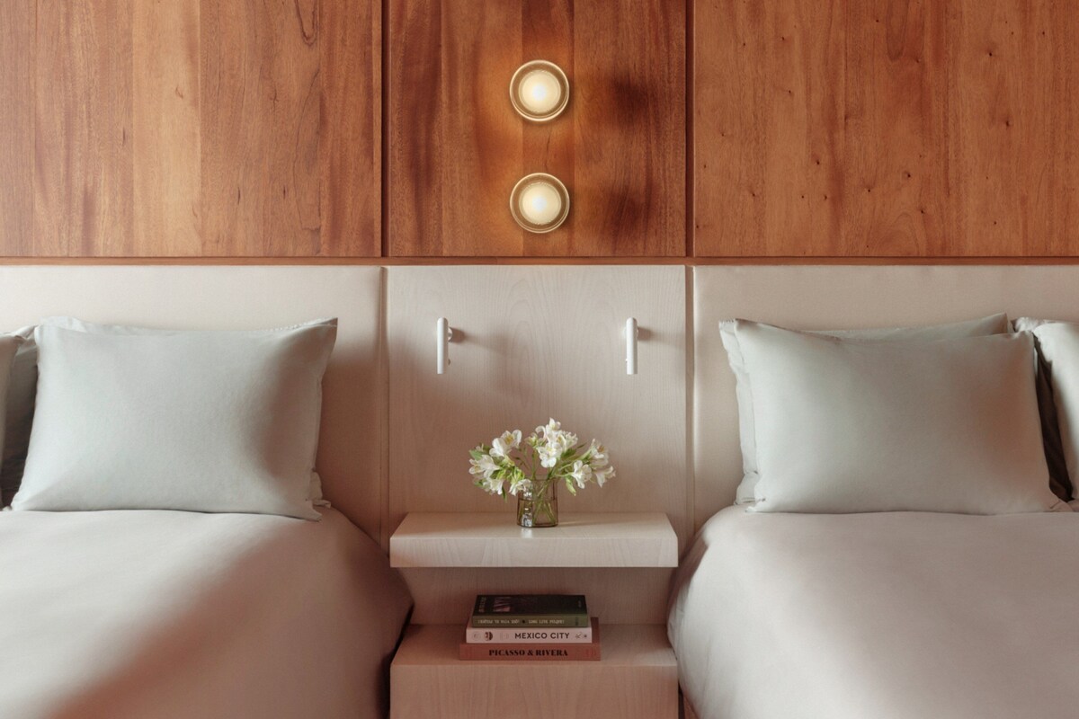 A hotel room with two beds, a nightstand with books, a vase with flowers, and wooden paneling on the wall.