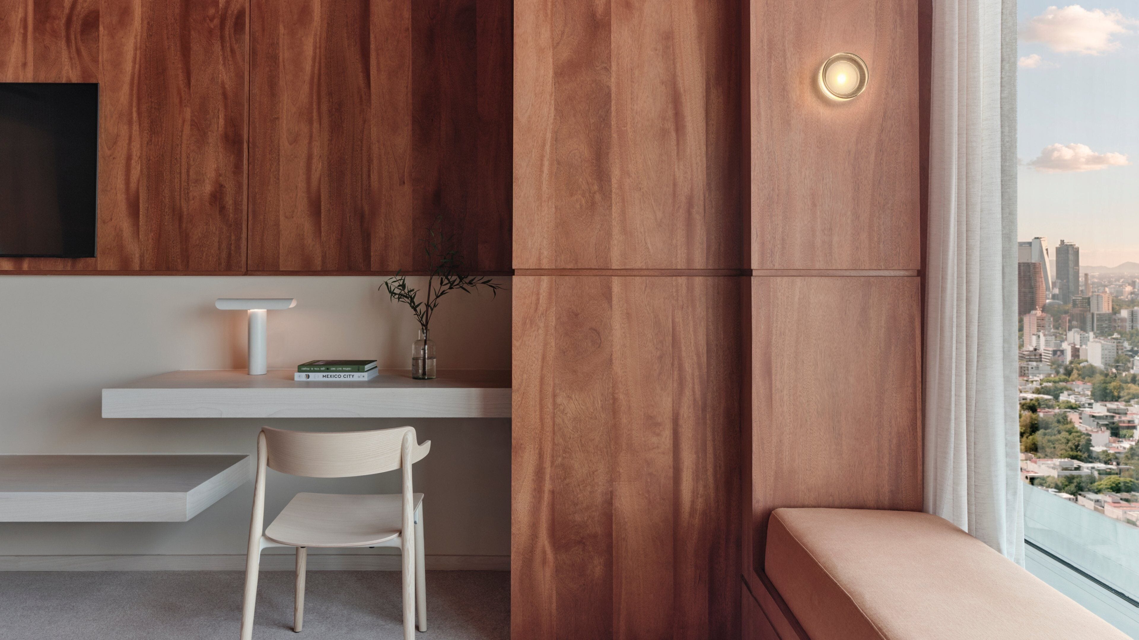 A modern room with a wooden accent wall, a white desk, a chair, a small table with a vase, and a view of a cityscape through a window.
