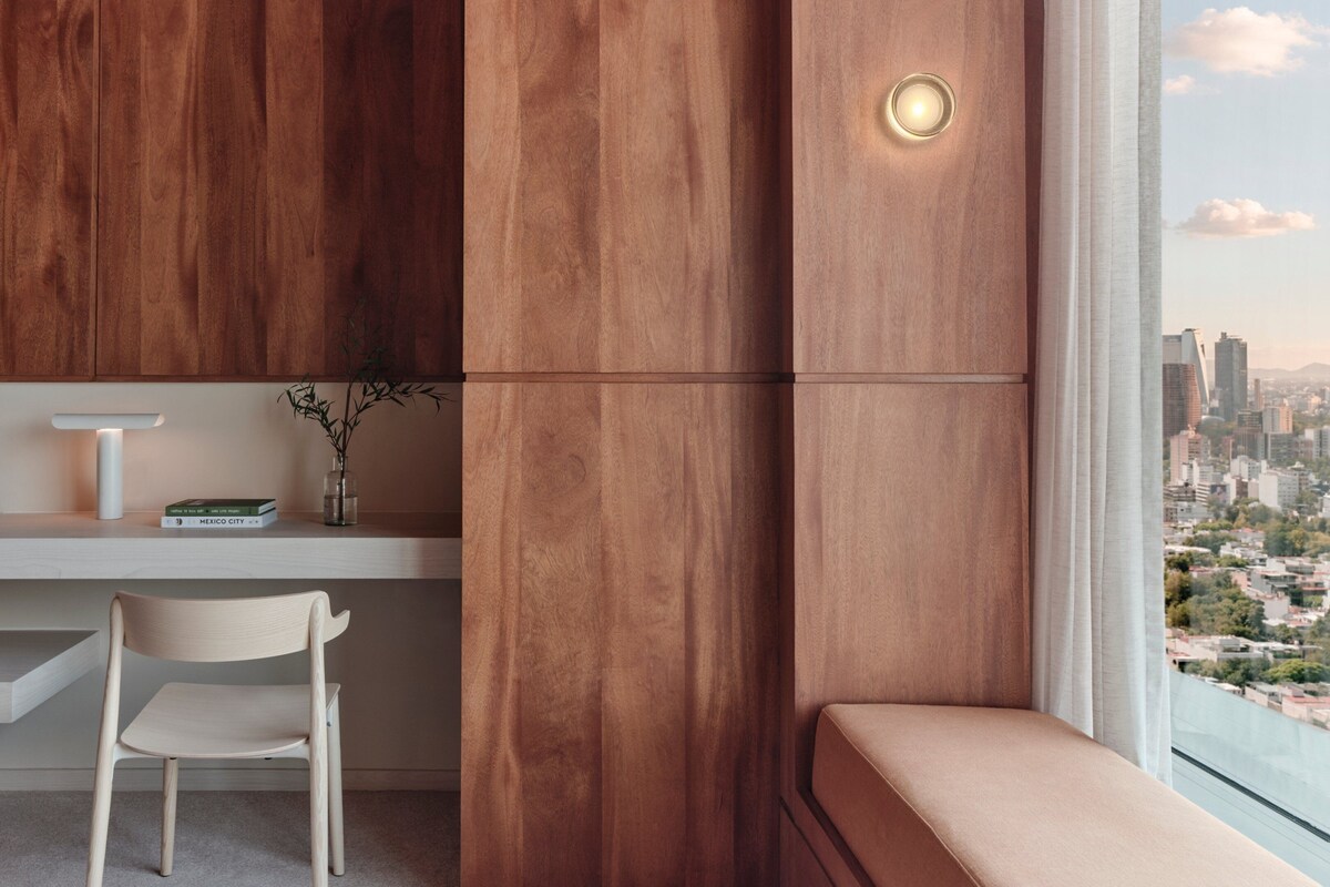 A modern room with a wooden accent wall, a white desk, a chair, a small table with a vase, and a view of a cityscape through a window.