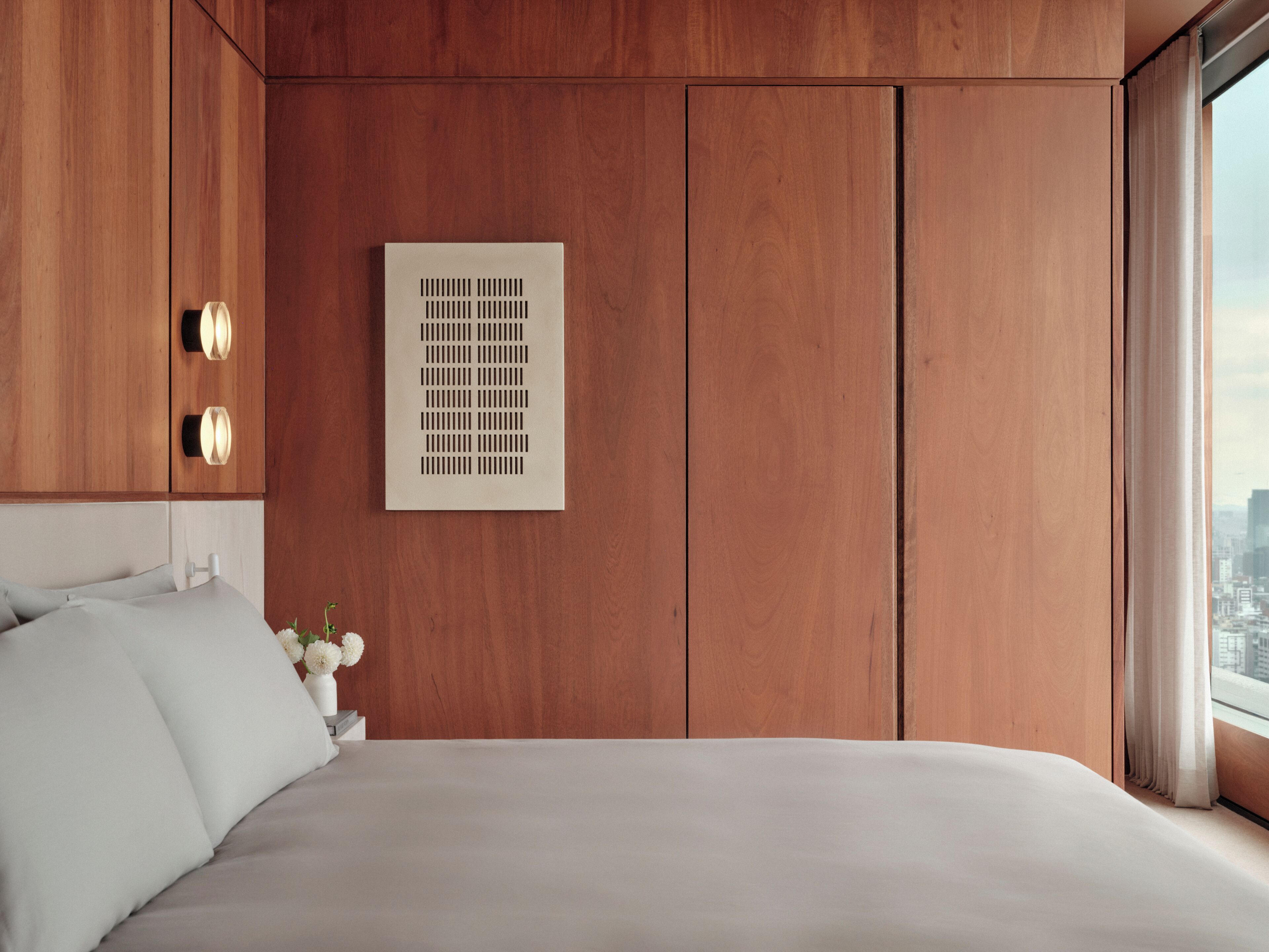 A bedroom with a bed, a wall-mounted air vent, and a window with curtains.