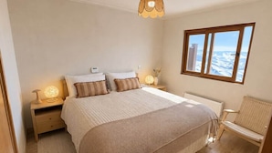 A bedroom with a large bed, bedside lamps, a view of the mountains through a window, and a wicker chair. - kery’s sweet home in valle nevado (Lo Barnechea)