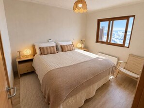 Un dormitorio con una cama grande, lámparas de noche, vistas a la montaña a través de una ventana y una silla de mimbre.