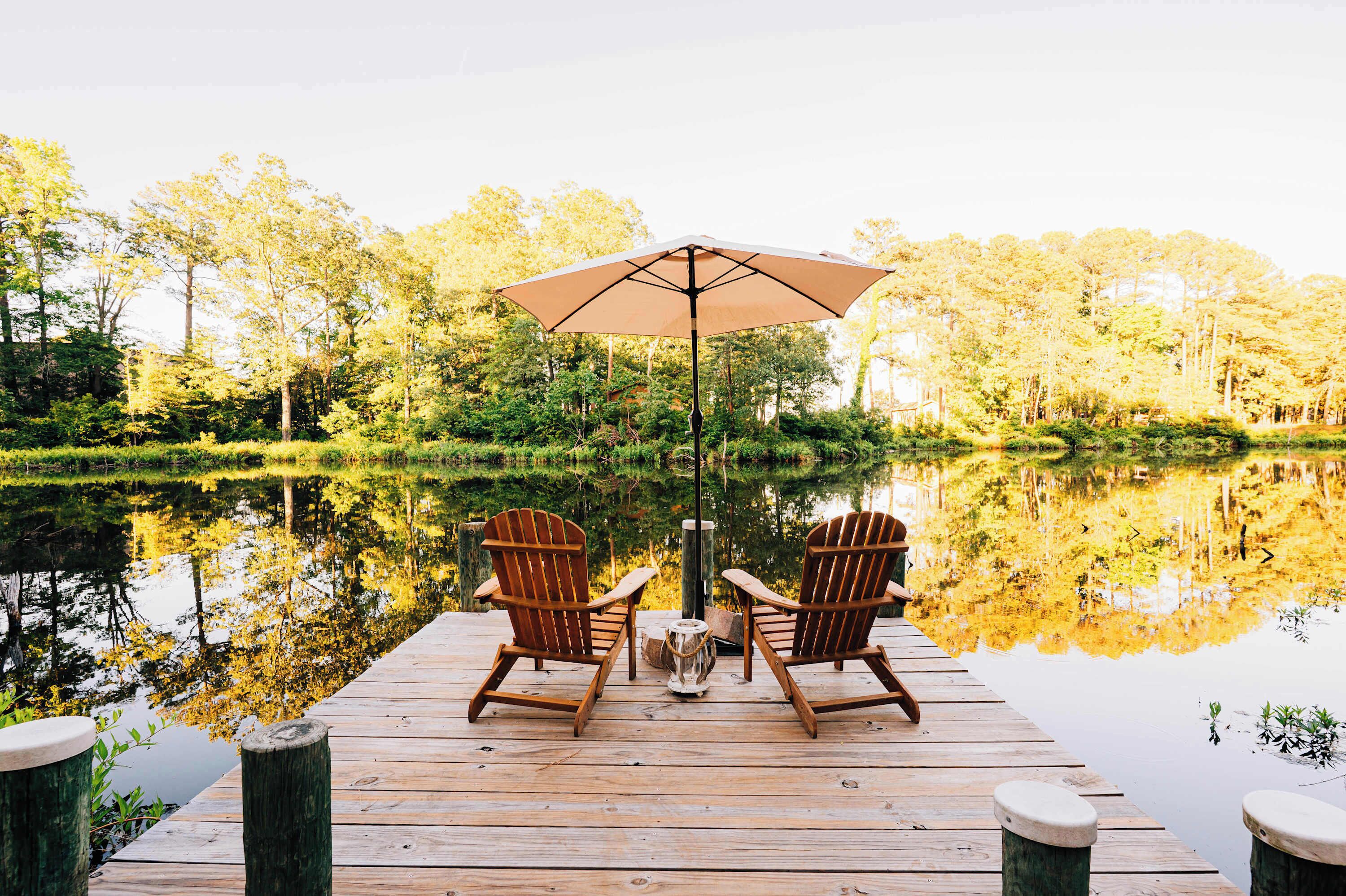 A wooden dock with two Adirondack chairs and a large umbrella, overlooking a calm lake surrounded by trees.