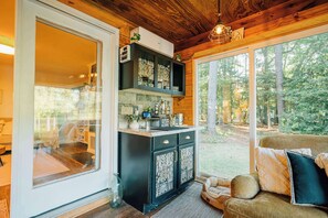 Ein gemütliches Blockhaus-Interieur mit Holzbalkendecke, einer Küchenzeile inklusive Kochfeld und einem großen Fenster mit Blick auf den Wald.