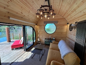 Ein gemütliches Blockhaushaus-Interieur mit Holzbalkendecke, einem Rundfenster, einer Couch, einem kleinen Tisch und Blick auf eine Holzterrasse draußen.